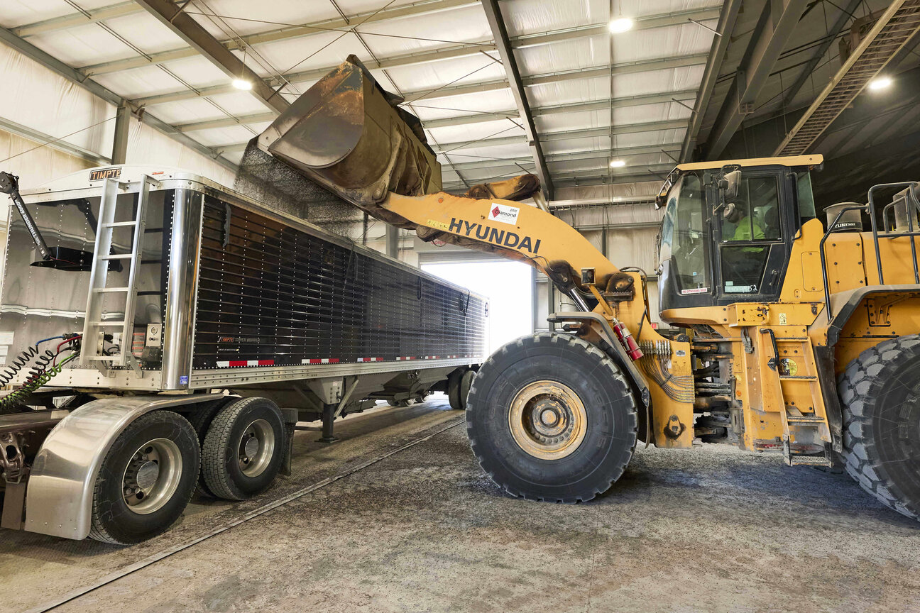 A large yellow loader is positioned beside a semi-truck, using its bucket to load material into the truck's trailer. The scene is set indoors, within a warehouse-like environment, with high ceilings and visible structural elements.