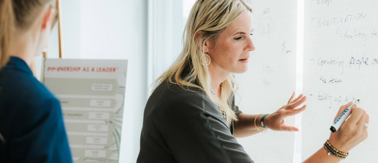 A woman writes on a whiteboard with a marker, sharing ideas during a discussion. Another person is partially visible in the background, indicating a collaborative environment. The focus is on the woman’s engagement and the writing on the board.