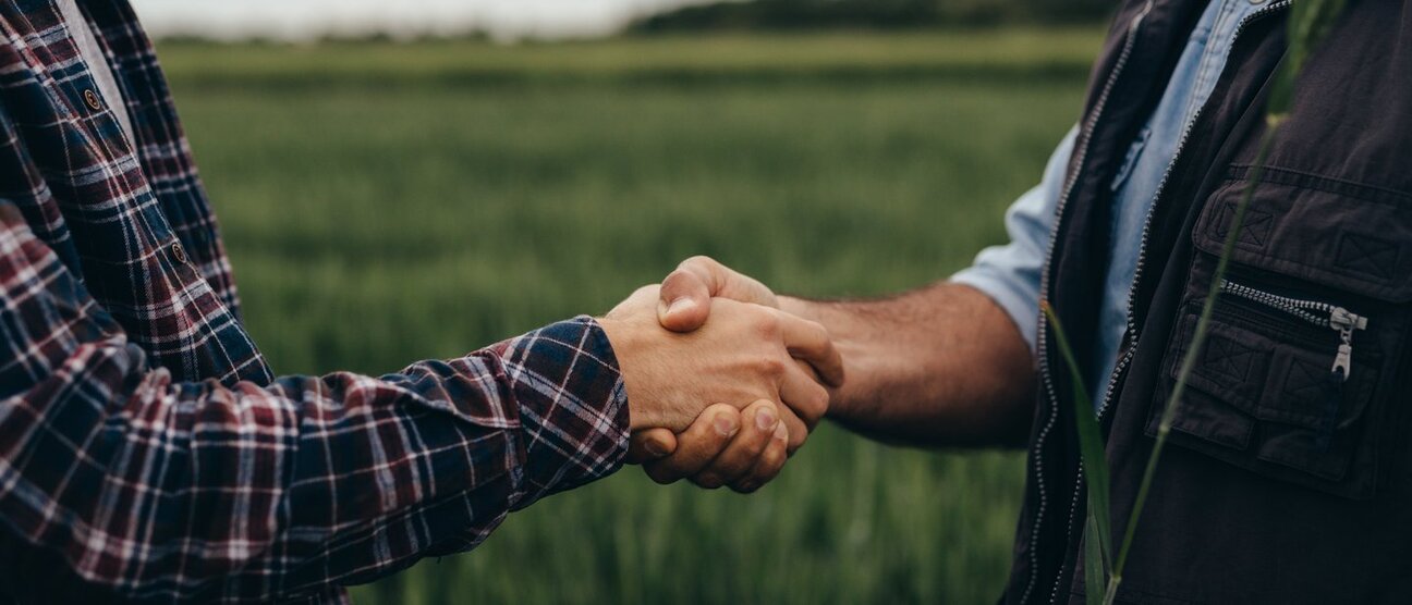 Two individuals are shaking hands in a green field, symbolizing partnership and collaboration. Their attire suggests an agricultural context. The handshake conveys trust and agreement between them.